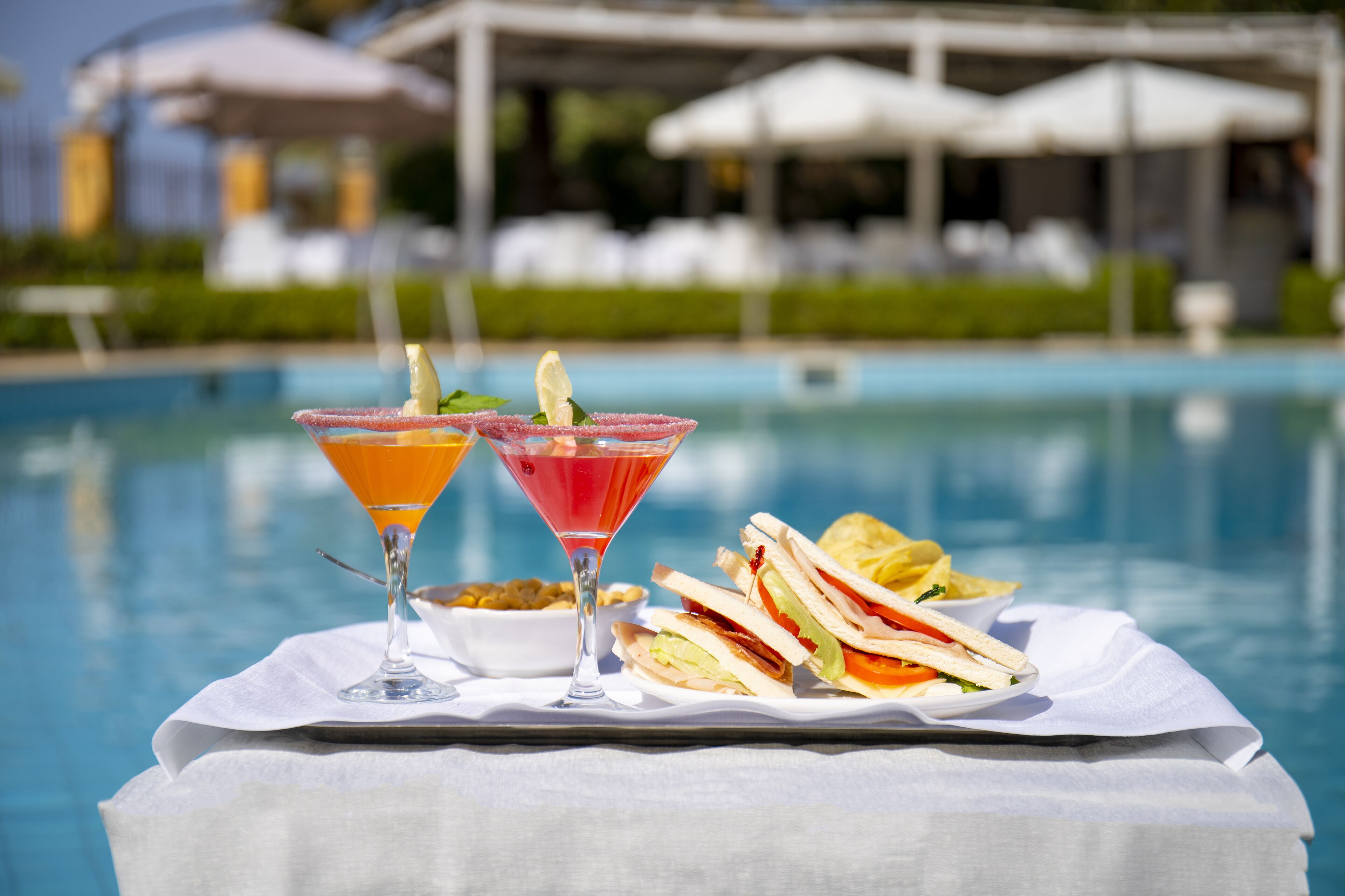 A poolside table with a sandwich and two drinks, a pool, and umbrellas in the background.