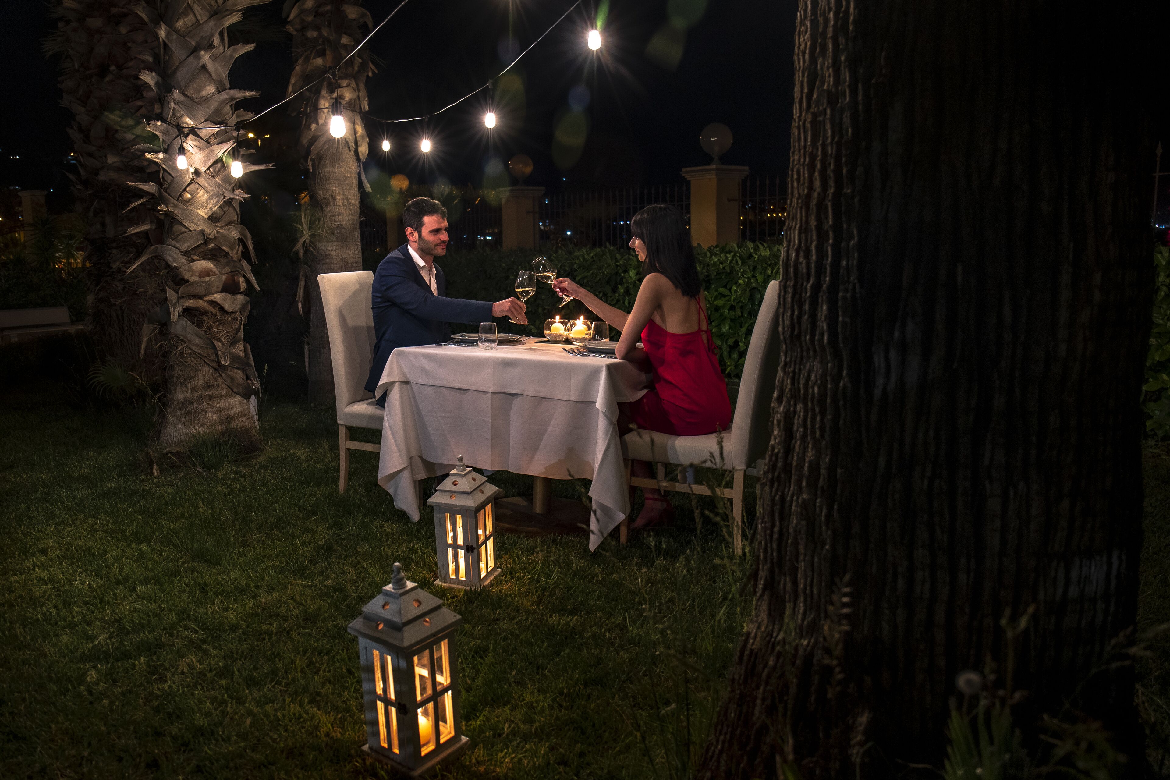 An outdoor dining area with a table set for two, surrounded by palm trees and string lights.