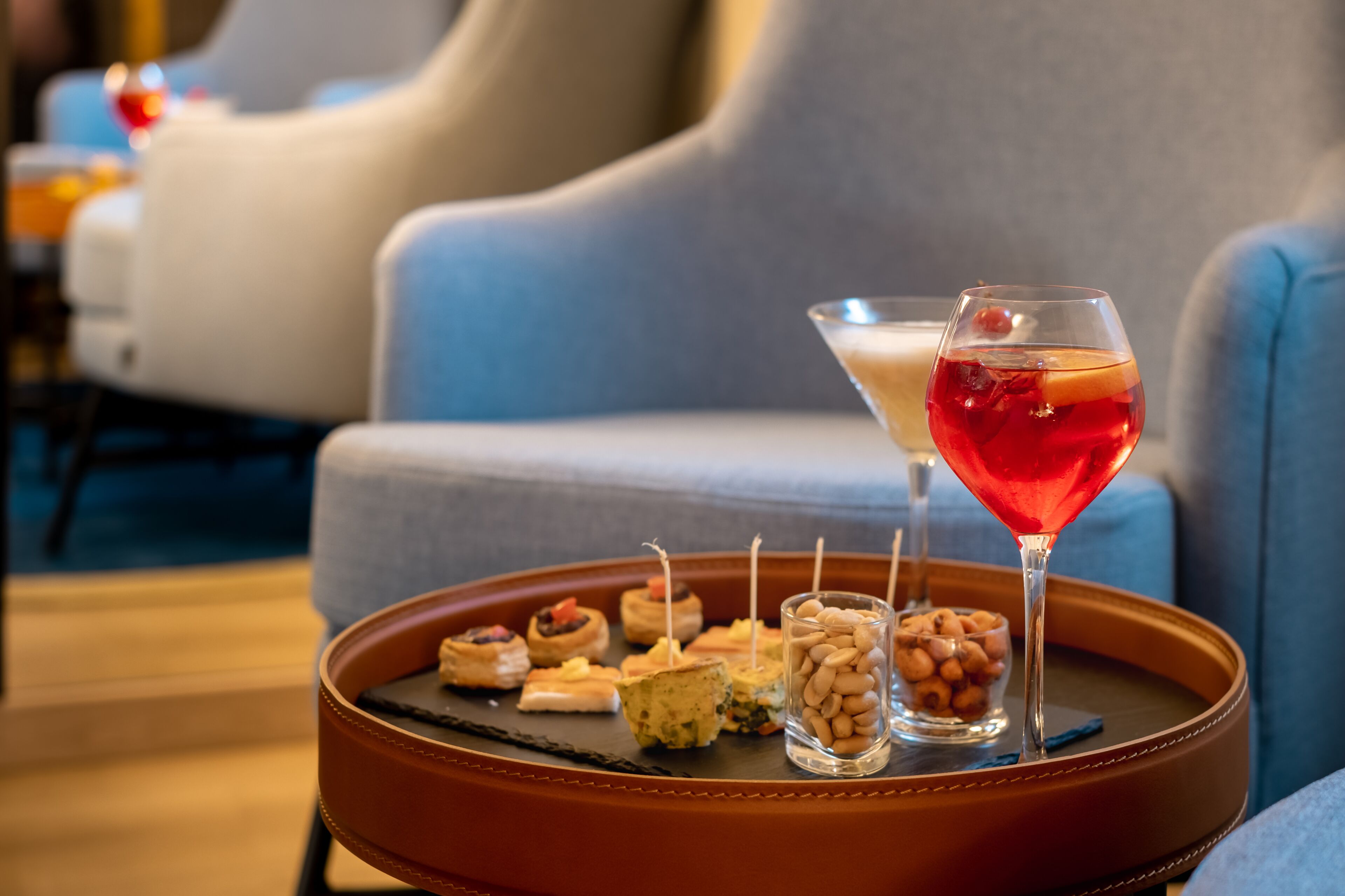 Uma bandeja com petiscos variados e um copo de martini sobre uma mesa em área de estar moderna, com sofás azuis.