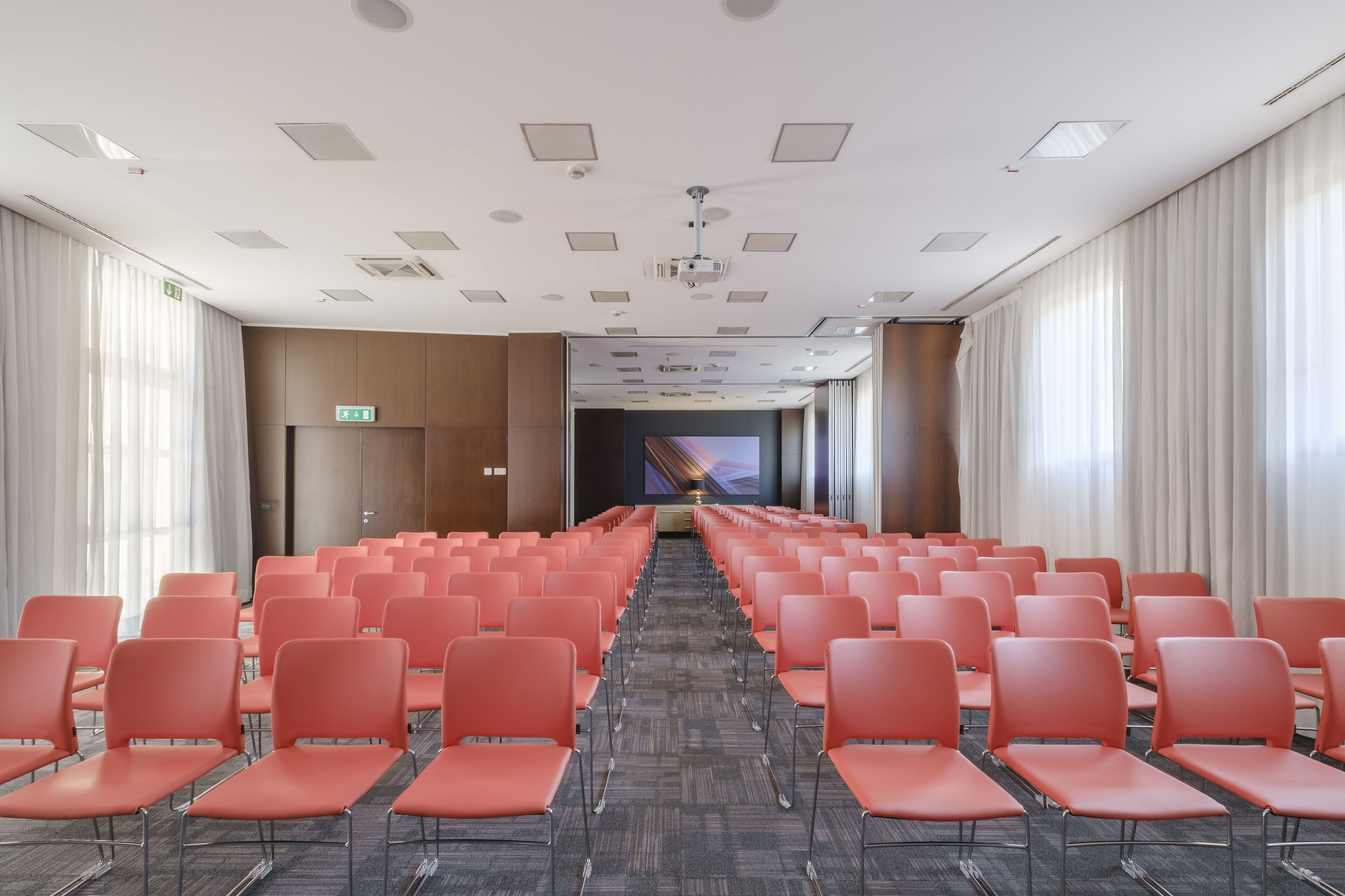 Un salón de conferencias con filas de sillas naranjas, una pantalla de proyector al frente y piso alfombrado.