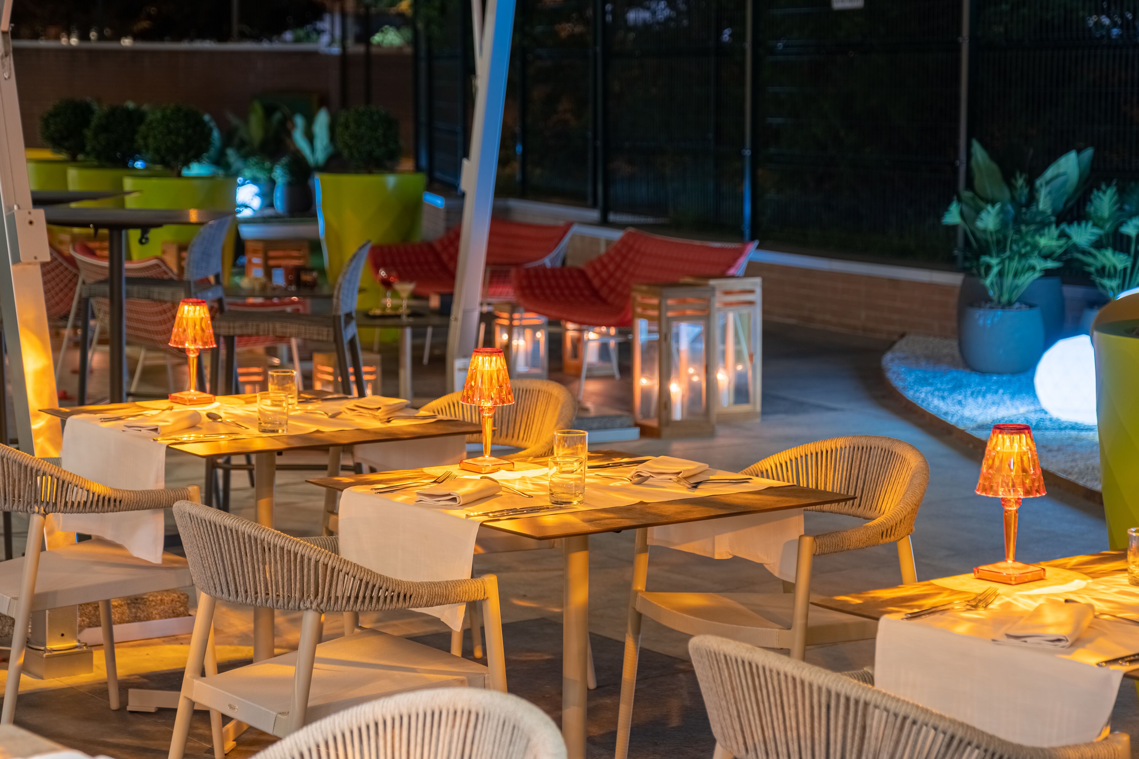 An outdoor dining area with tables and chairs, illuminated by table lamps.