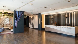 A modern hotel reception area with a marble counter, a receptionist, and a seating area with sofas and a plant.