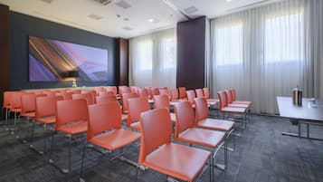 Une salle de conférence avec des rangées de chaises orange, un grand écran et une table équipée d’un microphone.