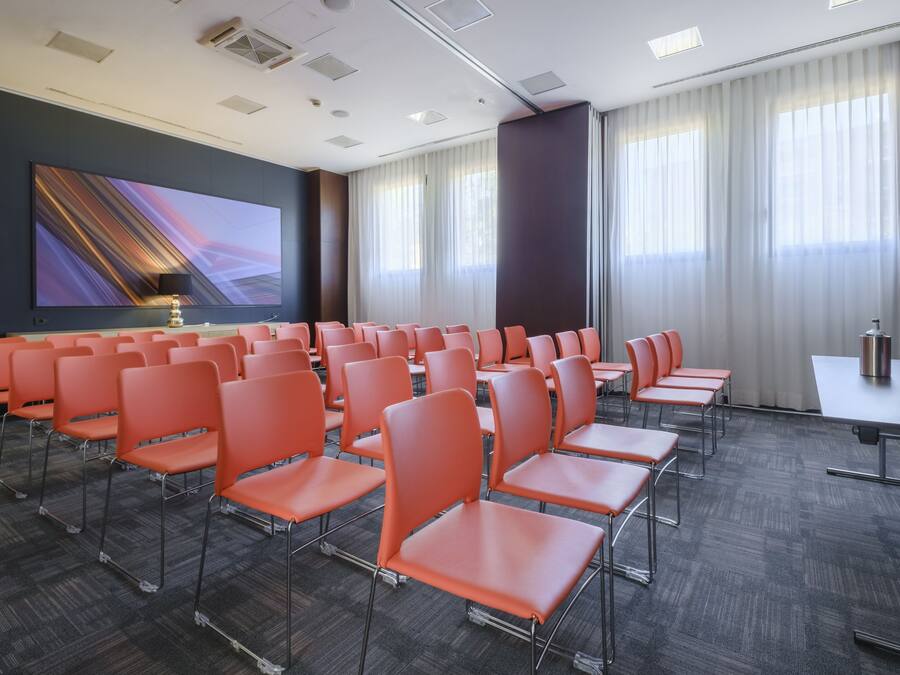 Une salle de conférence avec des rangées de chaises orange, un grand écran et une table équipée d’un microphone.
