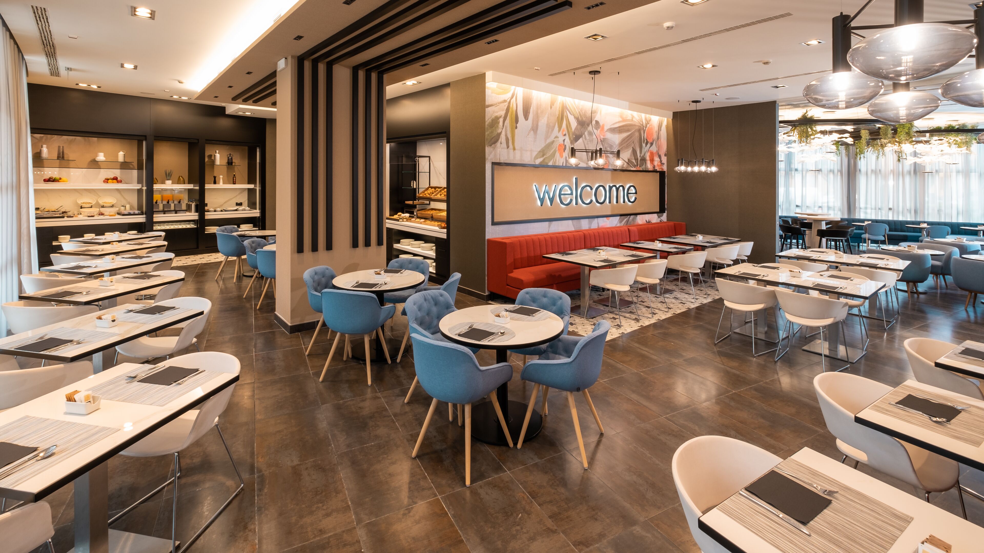 A modern restaurant with a 'welcome' sign, blue chairs, and a variety of seating options.