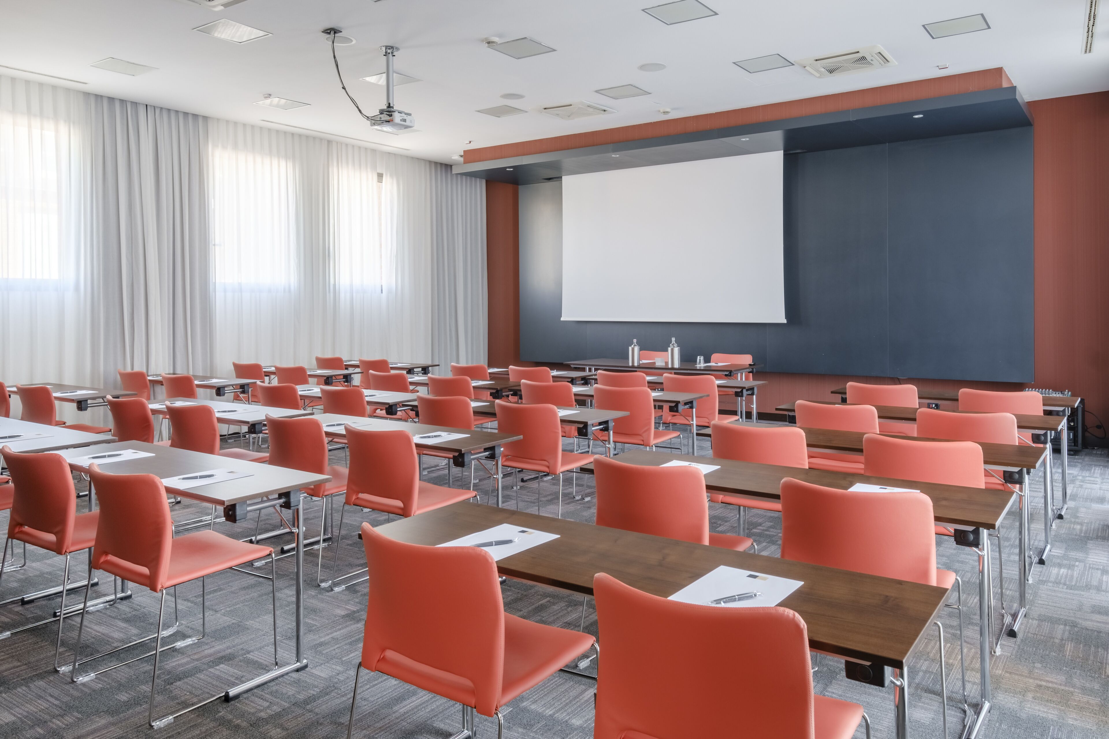 A conference room with rows of orange chairs and desks, a large projection screen, and a whiteboard.