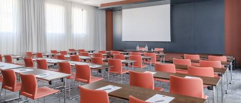 A conference room with rows of orange chairs and desks, a large projection screen, and a whiteboard.