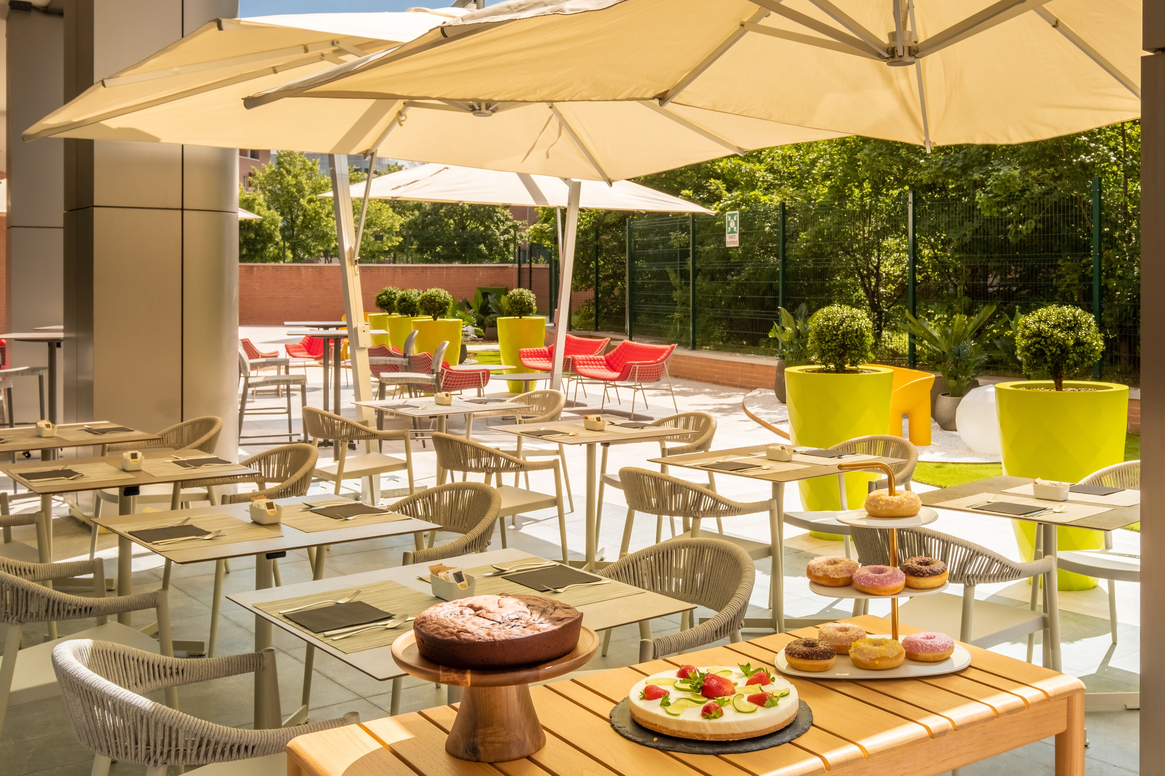 An outdoor café with tables and chairs under umbrellas, surrounded by greenery and a brick wall.