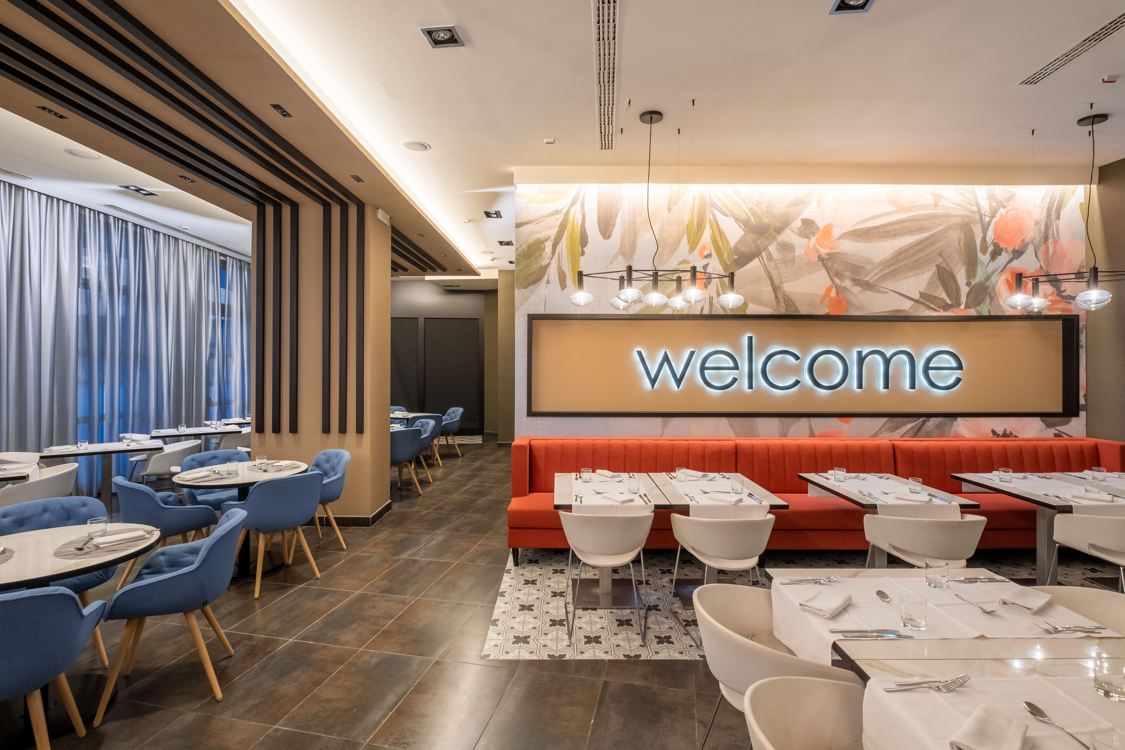 Een modern restaurant met een 'welcome'-bord, blauwe en witte stoelen en een rode zithoek. Tafels zijn gedekt met witte tafelkleden en bestek.