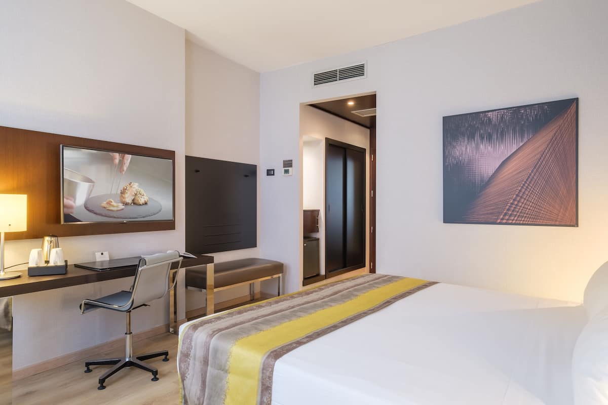A hotel room with a bed, desk, chair, and a large window.