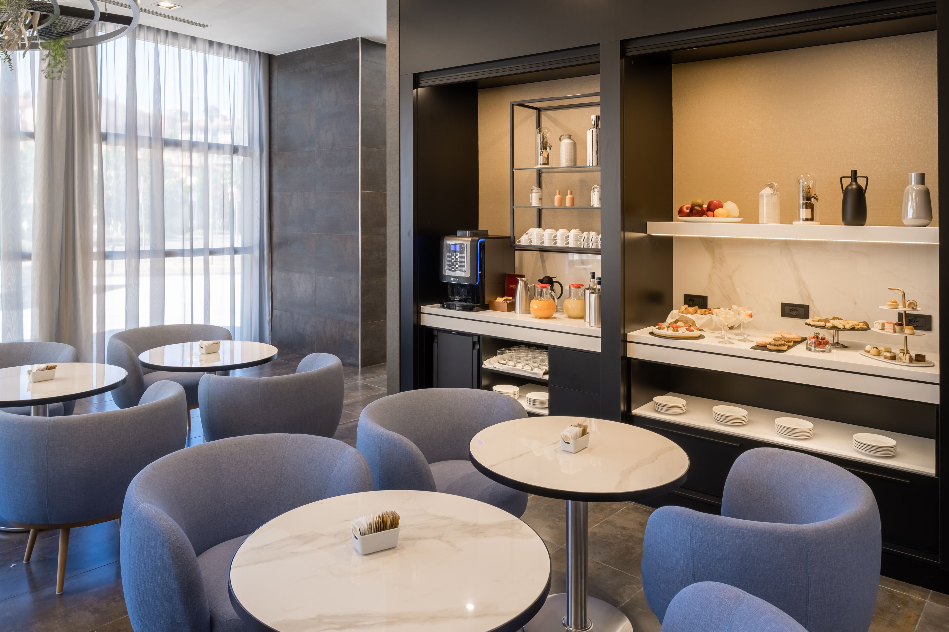 A modern hotel lobby with round tables and chairs, a coffee station, and a buffet area with various food items.