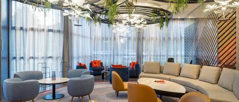 A modern lounge area with seating arrangements, a central table, and a ceiling with hanging plants and lighting fixtures.