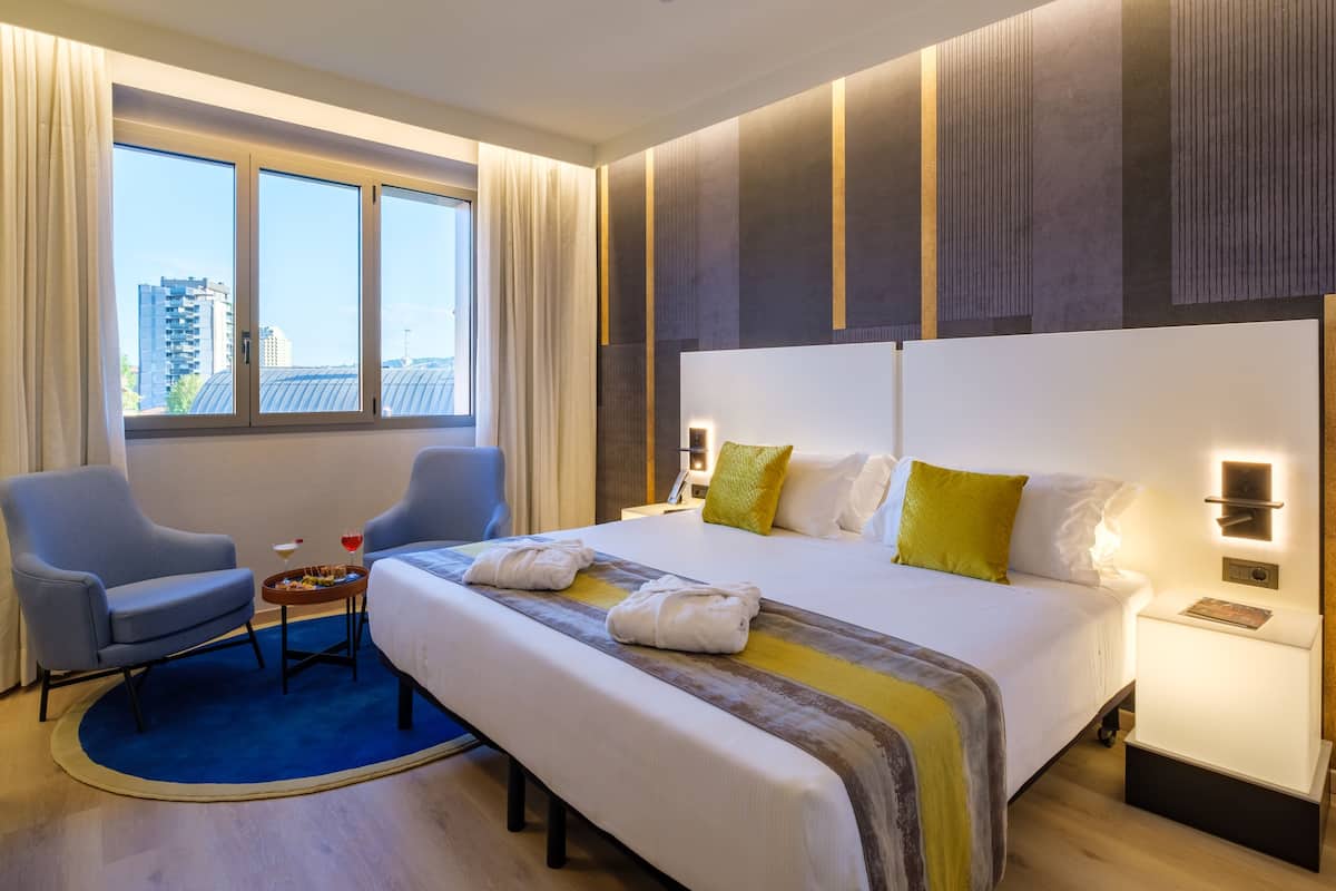 A hotel room with a large bed, two armchairs, a small table with a drink, and a view of buildings through the window.