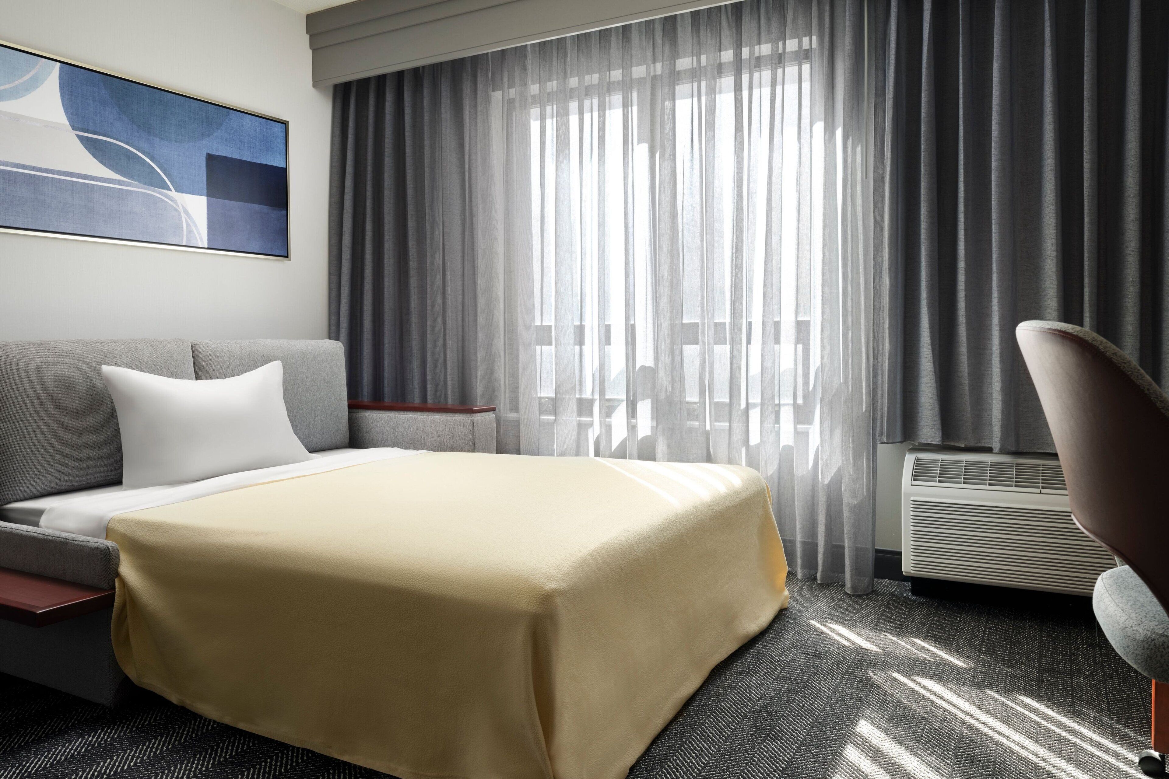 A hotel room with a bed, a sofa, a chair, a large window with curtains, and a wall-mounted air conditioner.