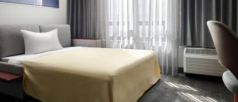 A hotel room with a bed, a sofa, a chair, a large window with curtains, and a wall-mounted air conditioner.
