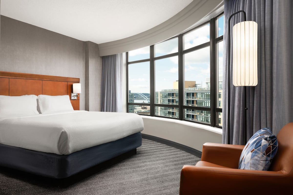 Una habitación de hotel con una cama grande, una silla, un ventanal con vistas a la ciudad y una lámpara moderna.