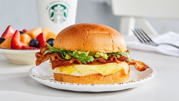 Een ontbijtsandwich met spek, avocado en ei op een broodje, een fruitschaal en een Starbucks-beker op de achtergrond.