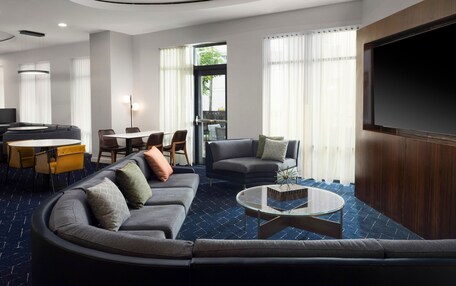 A modern hotel lobby with a curved sofa, a round coffee table, a TV, and a dining area with yellow chairs and a table.