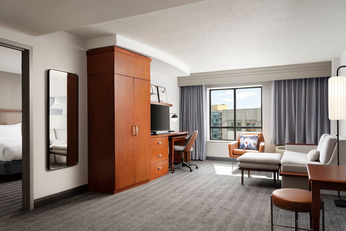 Habitación de hotel con cama, escritorio con silla, sofá, televisor y una ventana grande con cortinas.