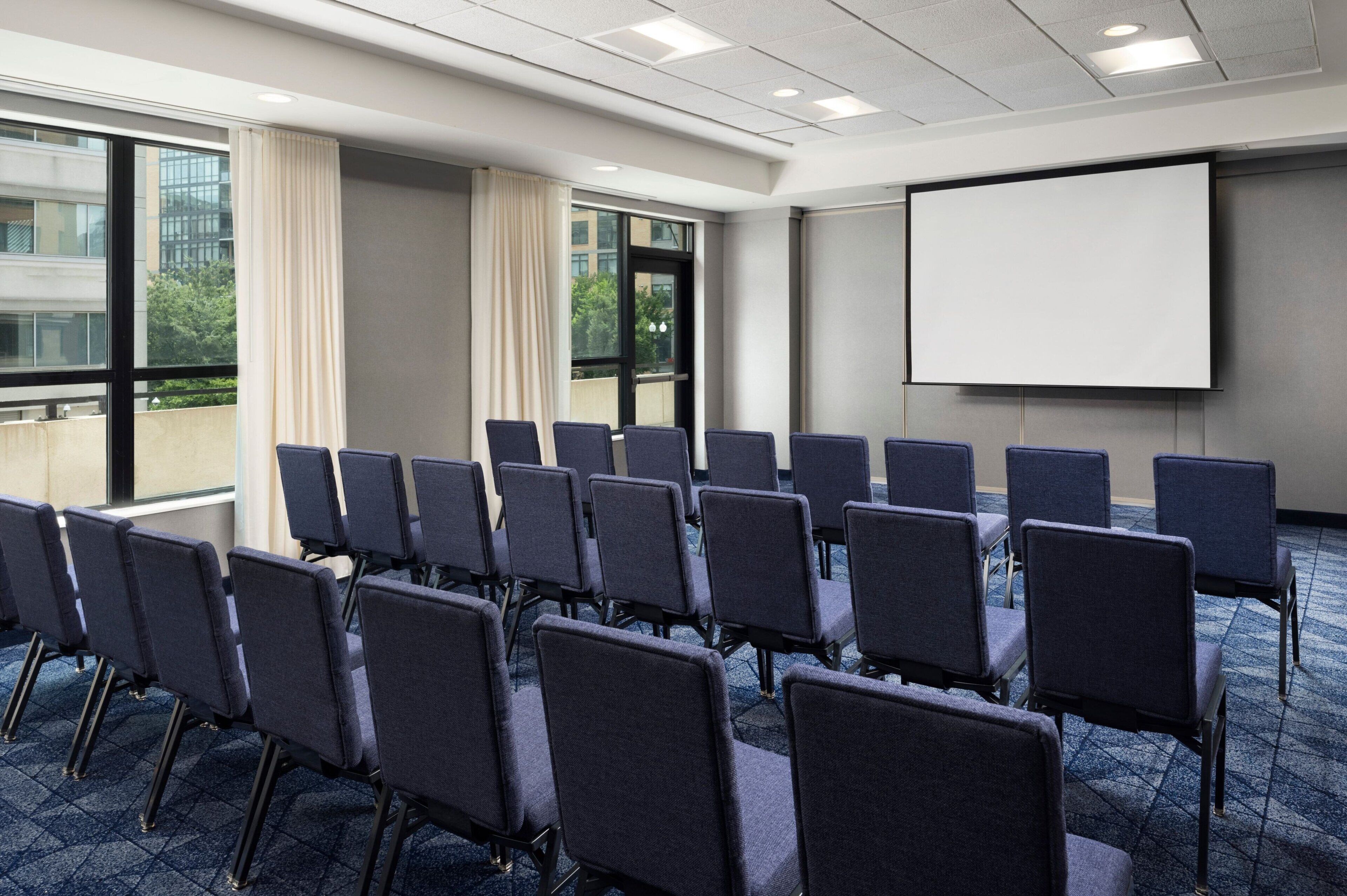 A conference room with rows of chairs, a projector screen, and large windows with curtains.