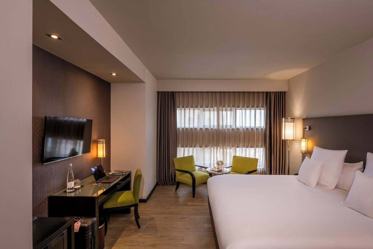 Ein Hotelzimmer mit Bett, Schreibtisch mit Laptop, Stuhl, Fernseher und einem Fenster mit Vorhängen.