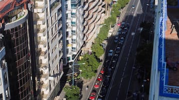 A multi-story building with balconies, a busy street with cars, and a clear view of the sea in the background.