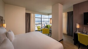 Habitación de hotel con una cama grande, un escritorio con silla, televisión y vistas a la ciudad a través de amplias ventanas.