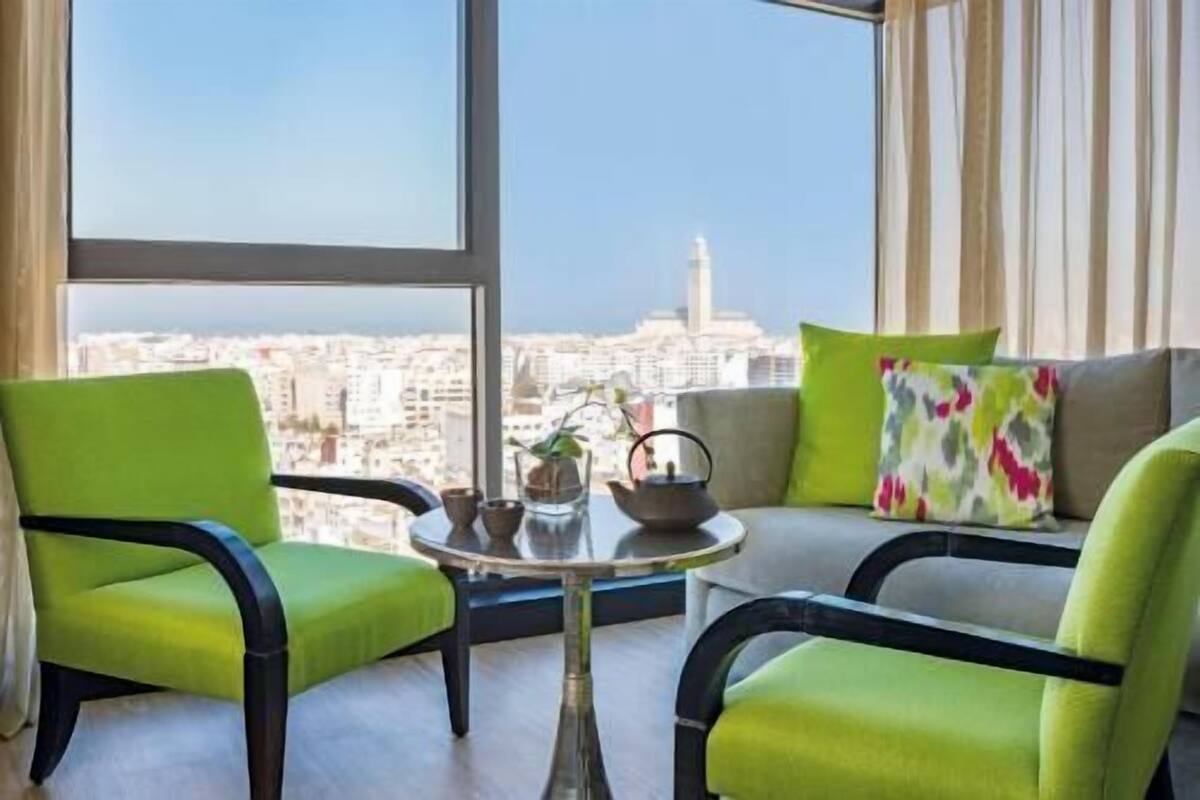 Ein Zimmer mit Blick auf eine Stadtlandschaft, zwei grüne Sessel, ein kleiner runder Tisch mit einer Teekanne und ein buntes Kissen auf einer Couch.