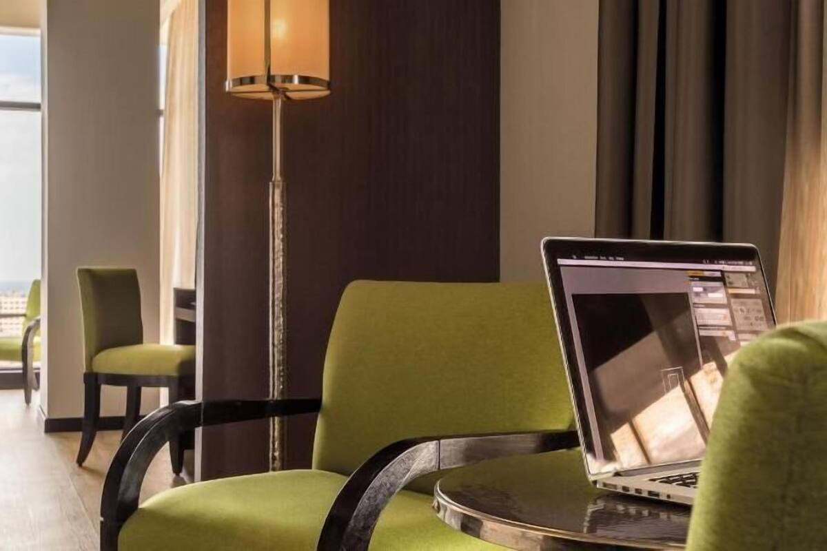 Ein Hotelzimmer mit Bett, Schreibtisch, Laptop, Stuhl, Stehlampe und Blick auf die Stadt durch das Fenster.