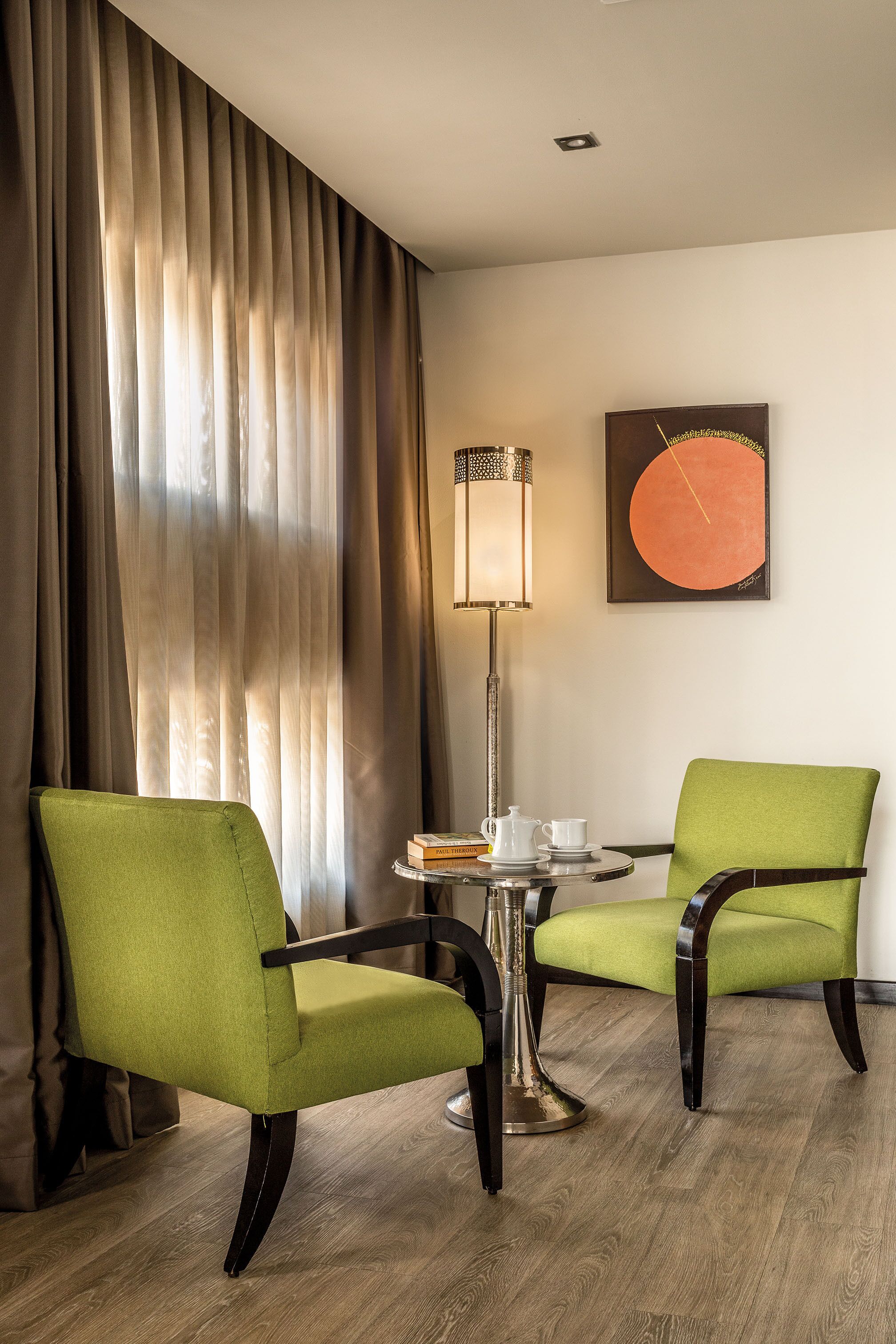 A hotel room with two green upholstered chairs, a small round table with a lamp, a white teapot, and a cup.