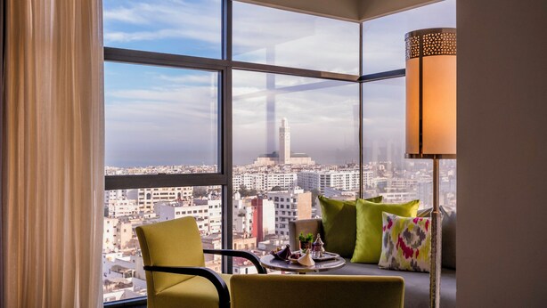 Habitación en un rascacielos con un ventanal grande que ofrece vistas a la ciudad, una mesa con una bandeja de objetos y una lámpara moderna.