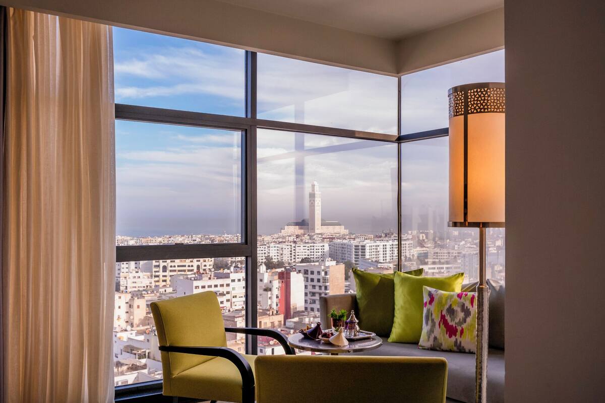 Ein Hotelzimmer in einem Hochhaus mit großem Fenster und Blick auf die Stadt, ein Tisch mit einer Auflage und eine moderne Lampe.