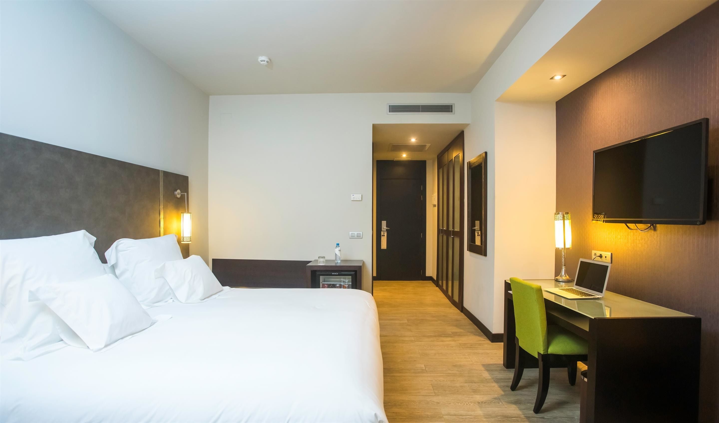A hotel room with a bed, desk, chair, TV, and a small table with a bottle and glass.