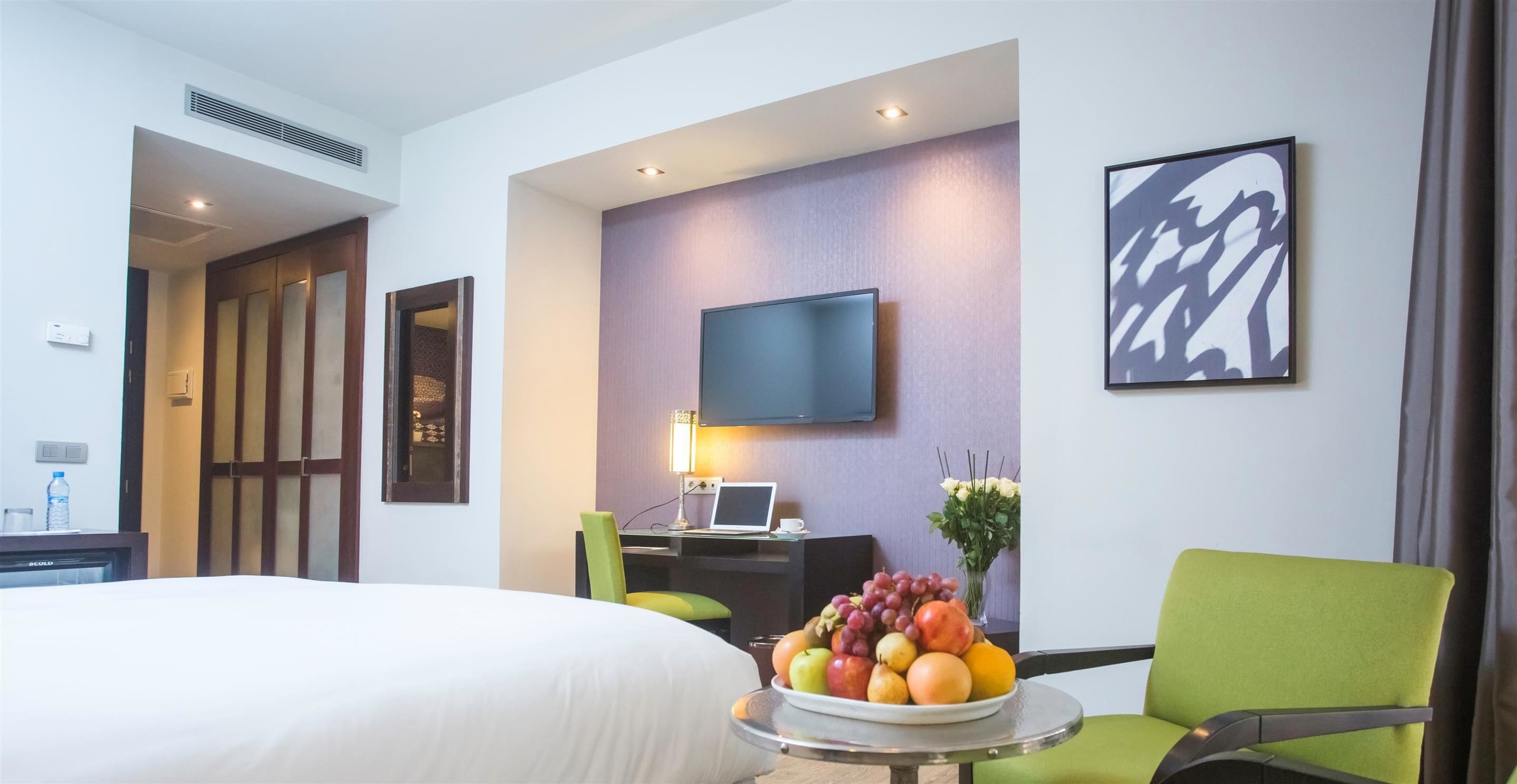 Une chambre d’hôtel avec un lit, un bureau avec un ordinateur portable, une télévision, un fauteuil vert, un panier de fruits et une œuvre d’art accrochée au mur.
