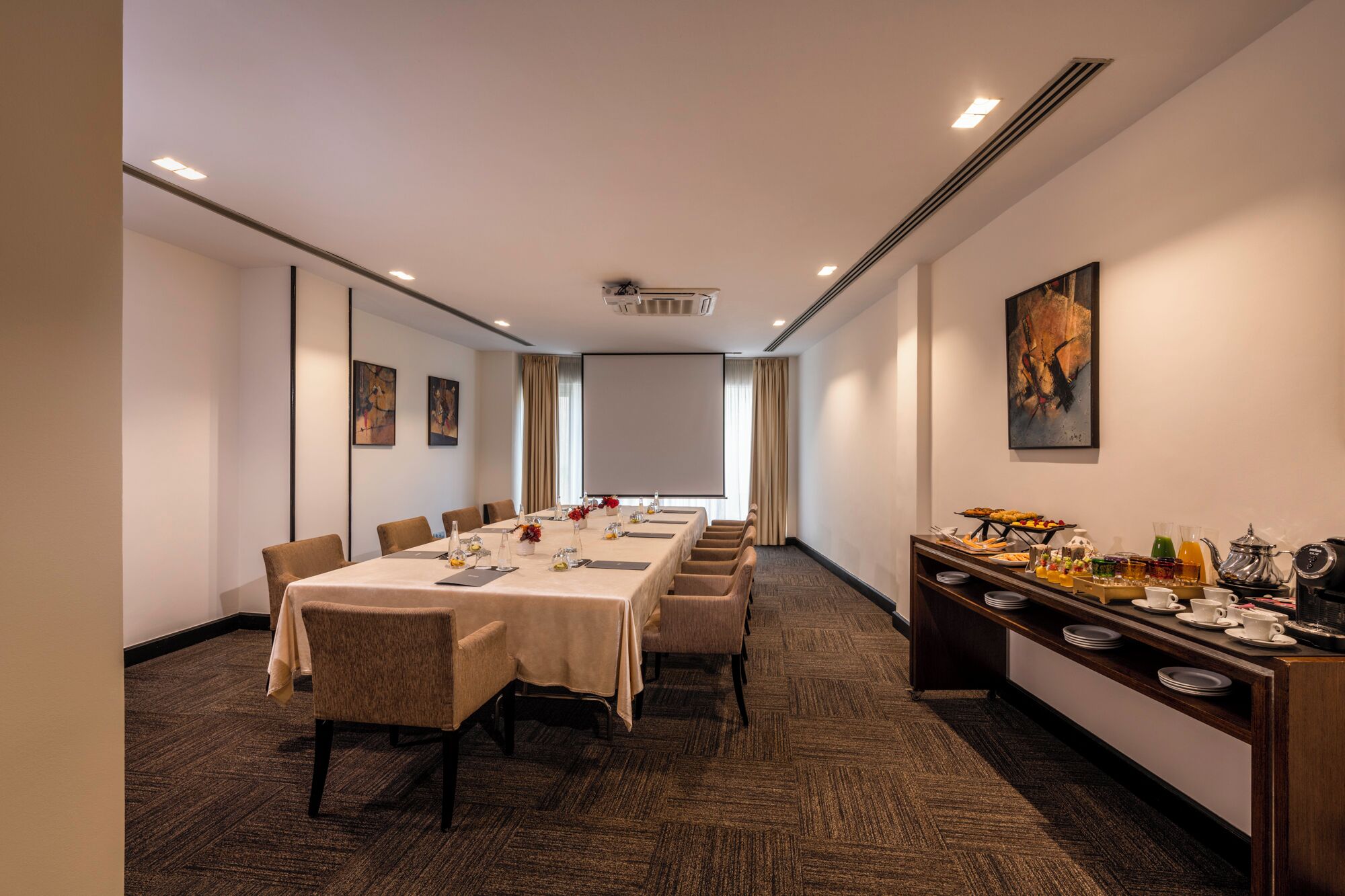 A conference room with a long table, chairs, a projector screen, and a buffet area with food and drinks.