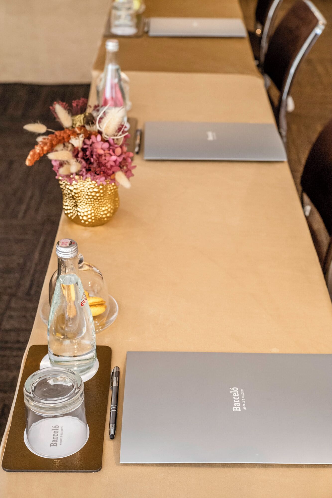 Una mesa de conferencias con computadoras portátiles, una botella de agua, un bolígrafo, un cuaderno y un jarrón decorativo con flores.