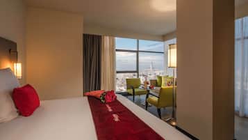 A hotel room with a large bed, a red runner, a city view, a sitting area with chairs, and a lamp.