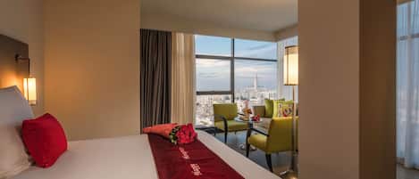 Habitación de hotel con una cama grande, un corredor rojo, vistas a la ciudad, una zona de estar con sillas y una lámpara.