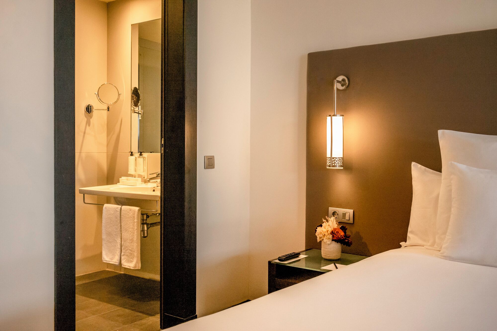 Ein Hotelzimmer mit einem Bett, einem Nachttisch mit Blumenvase, einer Wandleuchte und einem durchgehenden Türspalt sichtbaren Badezimmer.