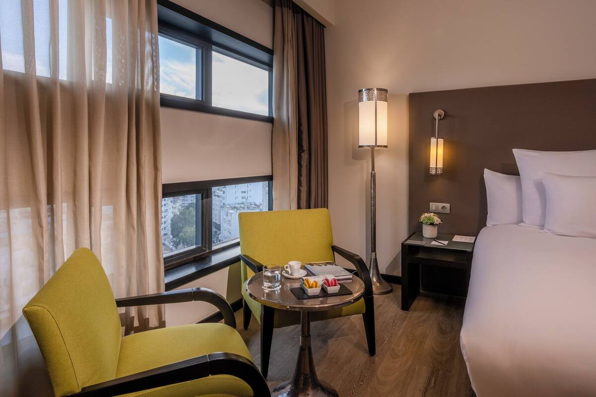 Ein Hotelzimmer mit Bett, zwei Stühlen, einem kleinen Tisch mit Tasse und Buch, einem Nachttisch mit Lampe und einem Fenster mit Vorhängen.