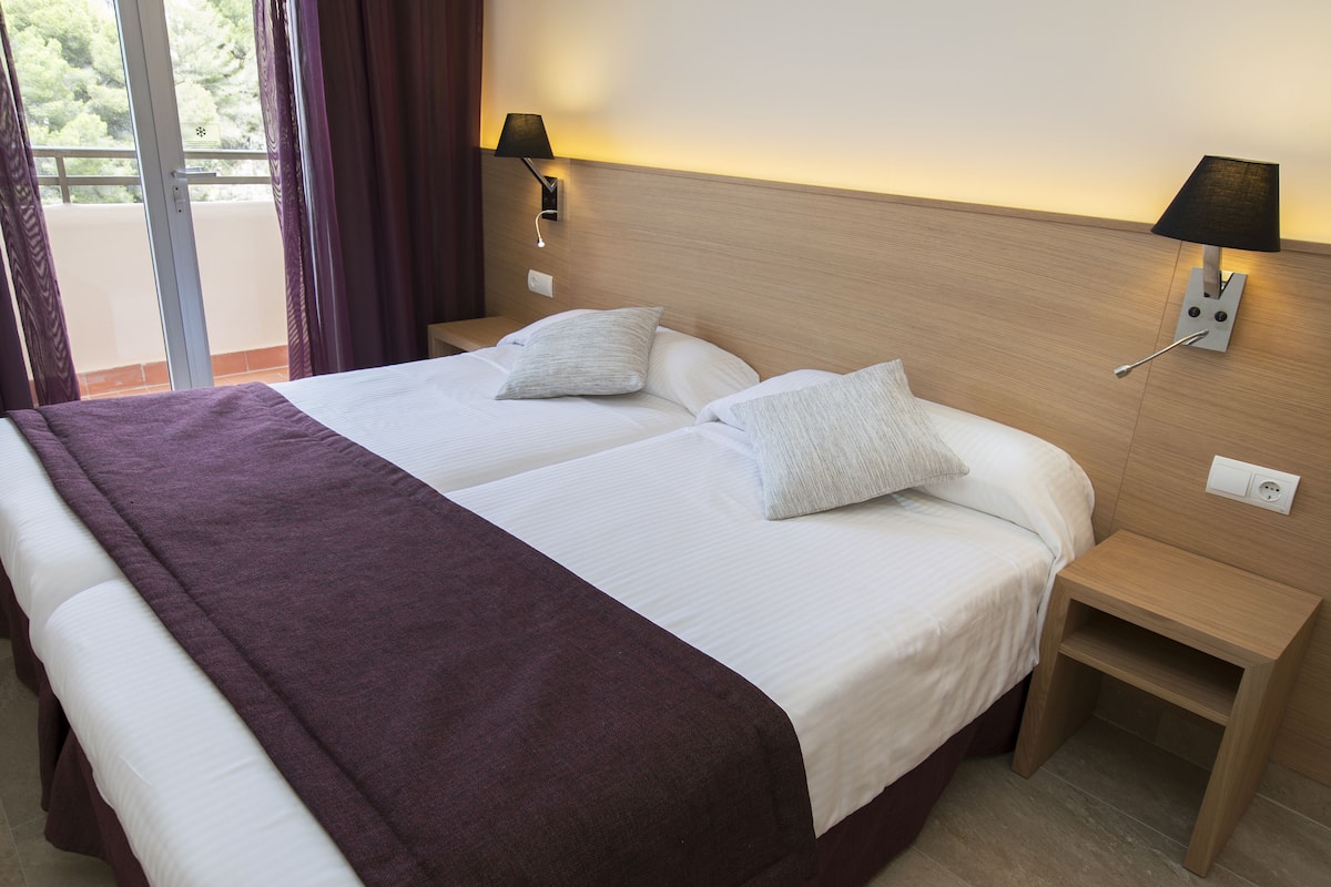 Ein Doppelbett mit einer violetten Tagesdecke, zwei Kissen und einem Nachttisch aus Holz mit einer Lampe.