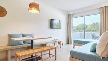 A modern hotel room with a large window, a flat-screen TV, a wooden bench, a small table, and a sofa with cushions.