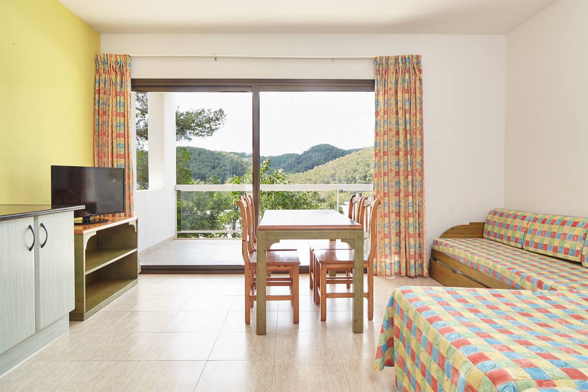 Ein Zimmer mit Esstisch, Stühlen, Fernseher und einem Bett mit gemusterten Bettbezügen.