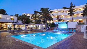 A swimming pool with lounge chairs and umbrellas, surrounded by palm trees and a multi-story building with balconies.