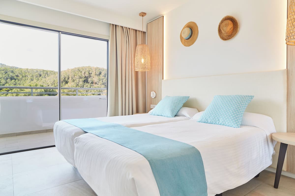 Ein Hotelzimmer mit zwei Betten, einem großen Fenster mit Blick auf Grünflächen, einer Hängelampe und zwei an der Wand befestigten Hüten.