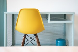 Una silla amarilla con patas de madera y un cubo de basura azul frente a un escritorio metálico azul claro con panel trasero de malla.