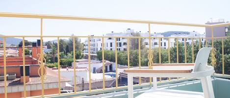 A balcony with a white chair and table, overlooking a residential area with white buildings and greenery.