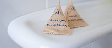 Deux produits de bain triangulaires, portant les mentions « GEL & SHAMPOO » et « GEL DE BAIN & SHAMPOO », sont posés sur une étagère blanche dans une salle de bain.