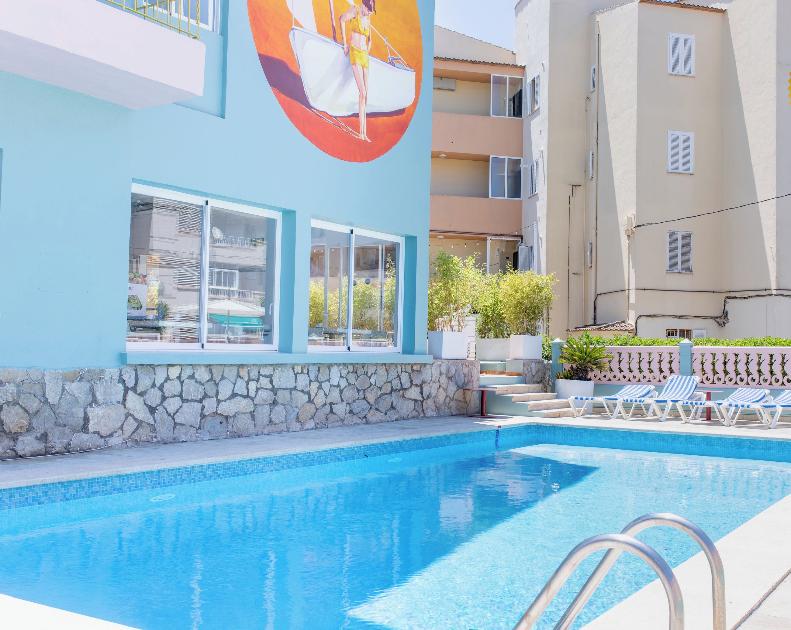 Une piscine avec des chaises longues, un mur en pierre et un bâtiment orné d’une fresque.