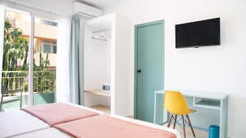 A hotel room with a bed, a desk, a chair, a TV, and a balcony with a view of buildings and plants.